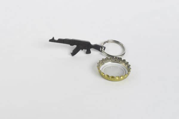 Kovaný prívesok - otvárak zbrane typu AK-47 uchopený na kľúčovom krúžku vedľa použitého korunkového uzáveru